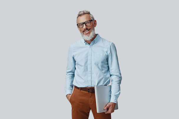 Confident mature man smiling and carrying laptop while standing against grey background - Foto, afbeelding