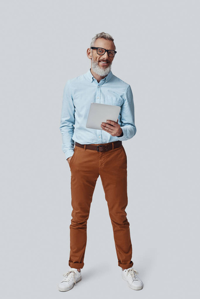 Full length of confident mature man looking at camera and using digital tablet while standing against grey background - Photo, Image