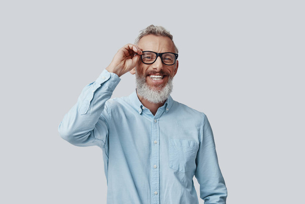 Happy mature man looking at camera and adjusting eyewear while standing against grey background - Foto, Imagem