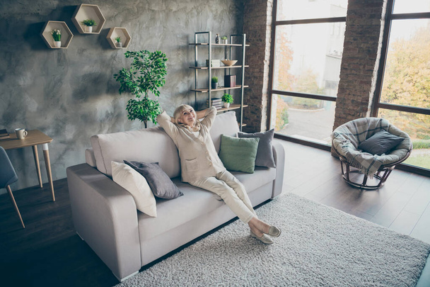 Full length profile photo of blond aged granny good mood hands behind head looking ceiling enjoy new house style design sitting comfort sofa divan living room indoors - 写真・画像