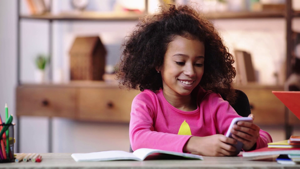 niño afroamericano sonriente usando un teléfono inteligente mientras hace la tarea
 - Metraje, vídeo