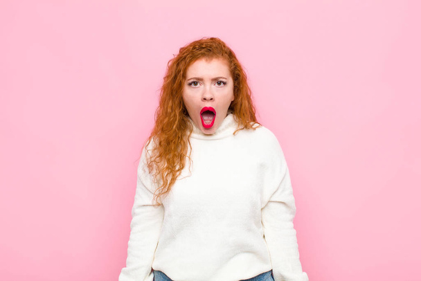 young red head woman looking shocked, angry, annoyed or disappointed, open mouthed and furious against pink wall - Foto, Imagem