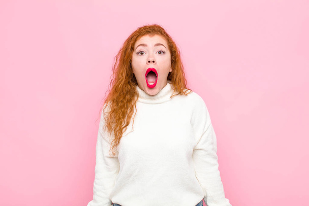 young red head woman feeling terrified and shocked, with mouth wide open in surprise against pink wall - Фото, изображение
