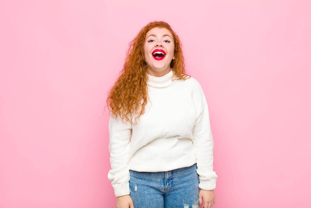 νεαρή κοκκινομάλλα γυναίκα αναζητούν χαρούμενος και ευχάριστα έκπληκτος, ενθουσιασμένος με μια συναρπαστική και σοκαρισμένη έκφραση κατά ροζ τοίχο - Φωτογραφία, εικόνα