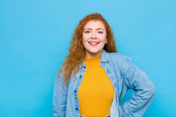 young red head woman smiling happily with a hand on hip and confident, positive, proud and friendly attitude against blue wall - Фото, изображение