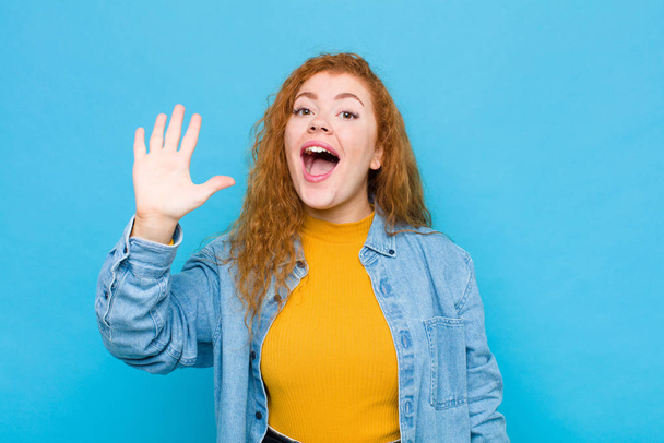 young red head woman smiling and looking friendly, showing number five or fifth with hand forward, counting down against blue wall - Фото, изображение