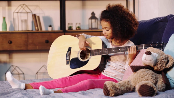 Afrykański amerykański dziecko gra na gitarze akustycznej w łóżku - Materiał filmowy, wideo