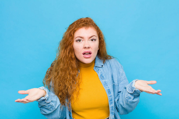 young red head woman feeling clueless and confused, not sure which choice or option to pick, wondering against blue wall - Photo, image