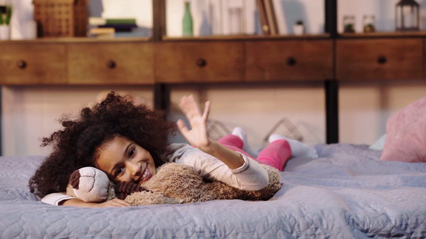 happy african american child waving hand while lying with teddy bear in bed - Footage, Video
