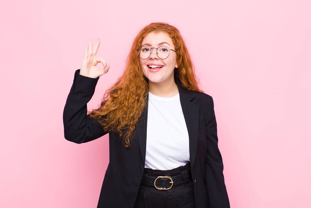 young red head woman feeling happy, relaxed and satisfied, showing approval with okay gesture, smiling against pink wall - Valokuva, kuva