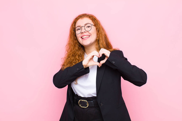 young red head woman smiling and feeling happy, cute, romantic and in love, making heart shape with both hands against pink wall - Foto, Imagen