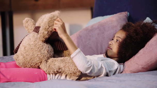 african american child playing with teddy bear while lying in bed - Filmagem, Vídeo