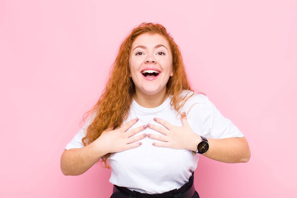 young red head woman looking happy, surprised, proud and excited, pointing to self against pink wall - Foto, imagen