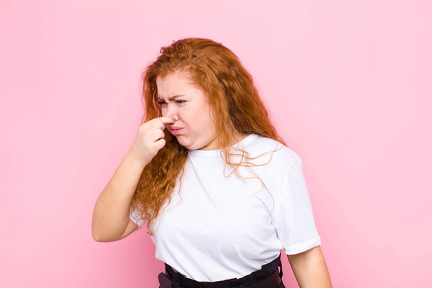 young red head woman feeling disgusted, holding nose to avoid smelling a foul and unpleasant stench against pink wall - Photo, image