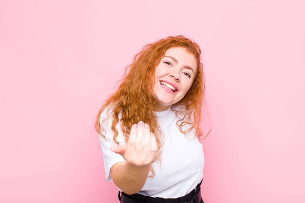νεαρή κοκκινομάλλα γυναίκα αισθάνεται ευτυχισμένη, επιτυχημένη και σίγουρη, αντιμετωπίζει μια πρόκληση και λέει να το φέρει σε! ή σας καλωσορίζουμε ενάντια σε ροζ τοίχο - Φωτογραφία, εικόνα