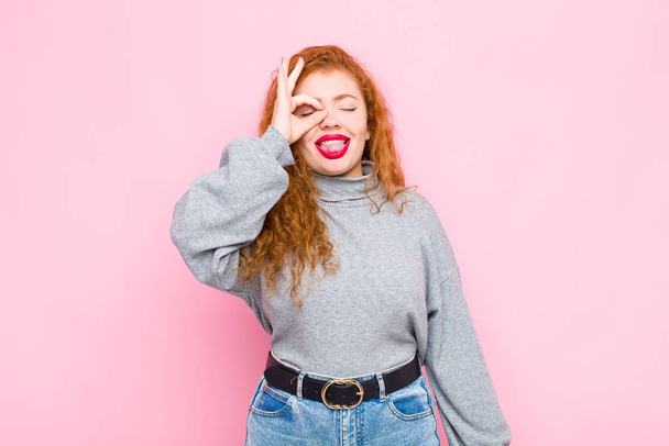 young red head woman smiling happily with funny face, joking and looking through peephole, spying on secrets against pink wall - Fotografie, Obrázek