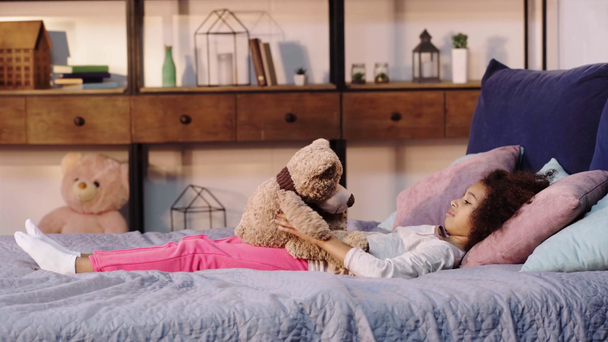 boční pohled na africké americké dítě hrát s medvídkem při ležení v posteli - Záběry, video