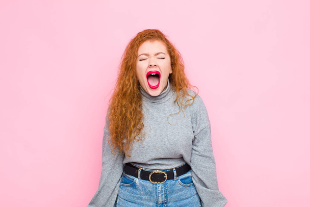 young red head woman shouting aggressively, looking very angry, frustrated, outraged or annoyed, screaming no against pink wall - 写真・画像