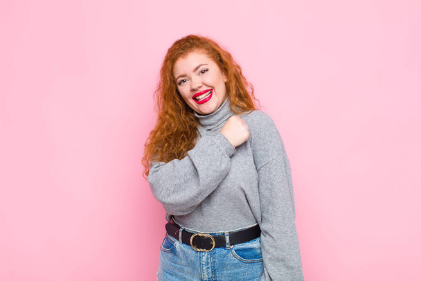 young red head woman feeling happy, positive and successful, motivated when facing a challenge or celebrating good results against pink wall - Photo, image