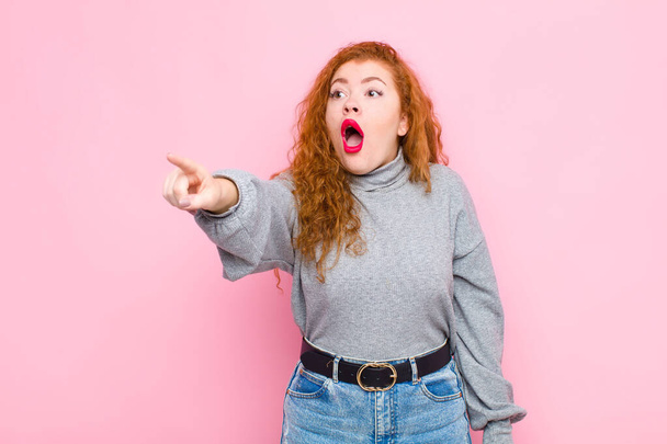 young red head woman feeling shocked and surprised, pointing and looking upwards in awe with amazed, open-mouthed look against pink wall - Photo, image