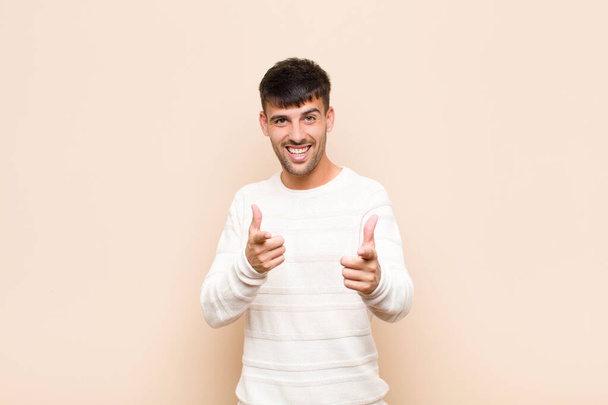 young handsome man feeling happy, cool, satisfied, relaxed and successful, pointing at camera, choosing you against warm wall - Foto, imagen