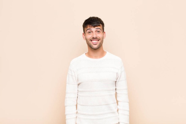young handsome man looking happy and goofy with a broad, fun, loony smile and eyes wide open against warm wall - Foto, Bild