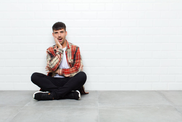 young handsome man open-mouthed in shock and disbelief, with hand on cheek and arm crossed, feeling stupefied and amazed sitting on cement floor - Foto, imagen