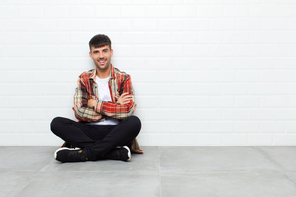 young handsome man looking like a happy, proud and satisfied achiever smiling with arms crossed sitting on cement floor - Foto, imagen