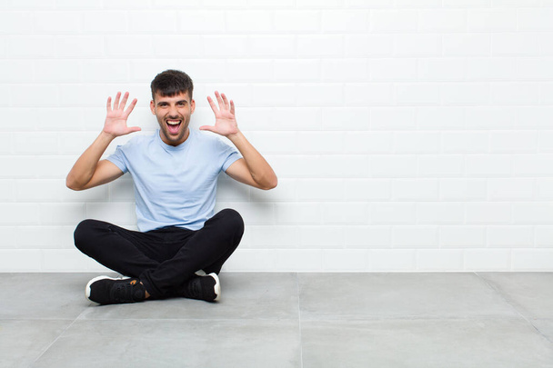 young handsome man screaming in panic or anger, shocked, terrified or furious, with hands next to head sitting on cement floor - Фото, изображение