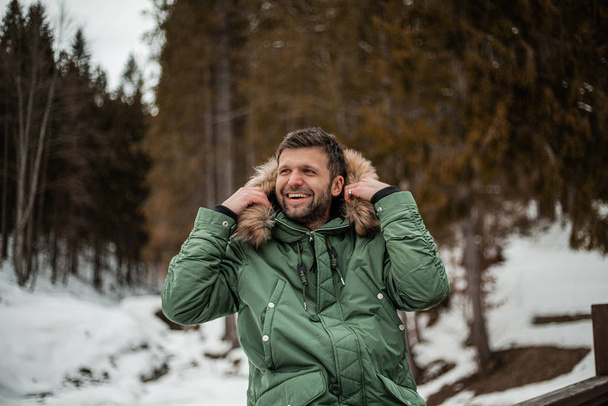 happy bearded man in warm coat sitting on bridge railing in snow-covered winter forest  - Photo, Image