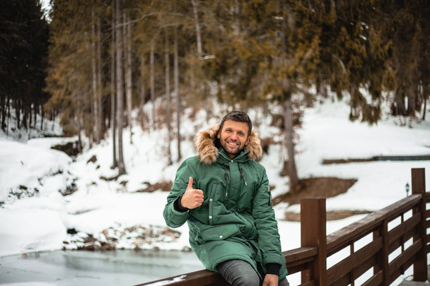 boldog szakállas férfi meleg kabátban mutatja hüvelykujját, és ül a hídon korláton hófödte téli erdőben  - Fotó, kép
