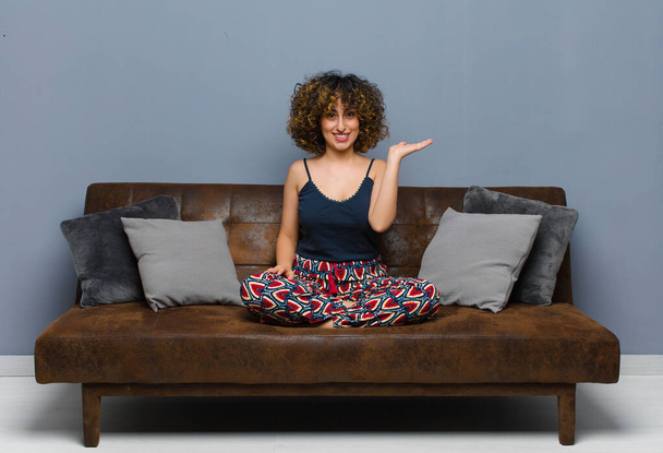 young pretty woman smiling, feeling confident, successful and happy, showing concept or idea on copy space on the side sitting on a sofa. - Photo, Image