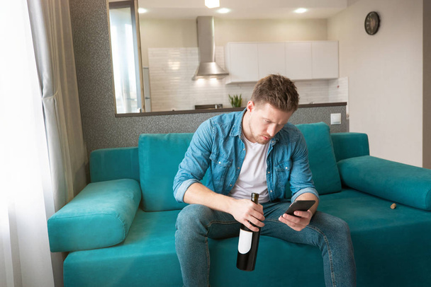 young handsome man sitting on the sofa with a bottle of wine texting message on his smartphone in the cozy living room looks nervous - Photo, Image