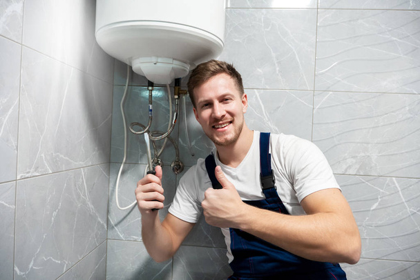 smiling man worker in uniform with screwdriver in one hand and showing like with another hand repairing boiler at home in toilette professional repair service - 写真・画像