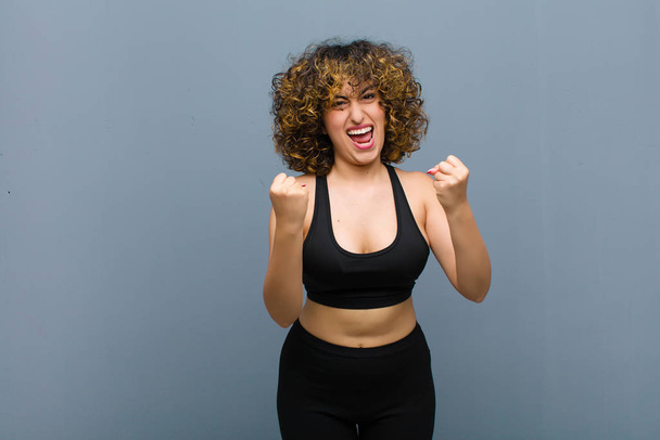 young sports woman shouting triumphantly, laughing and feeling happy and excited while celebrating success against gray wall - Фото, изображение