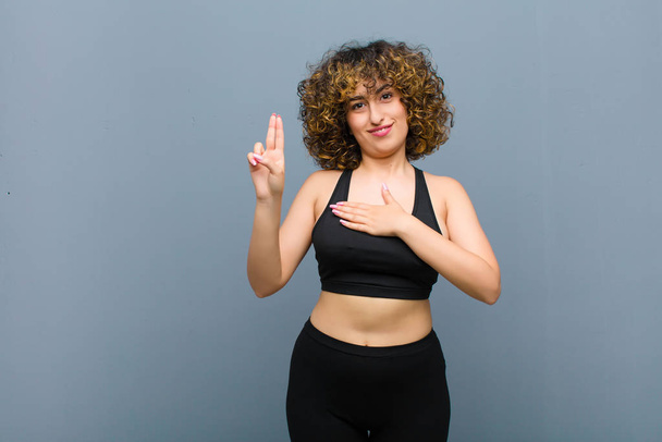 young sports woman looking happy, confident and trustworthy, smiling and showing victory sign, with a positive attitude against gray wall - Foto, afbeelding