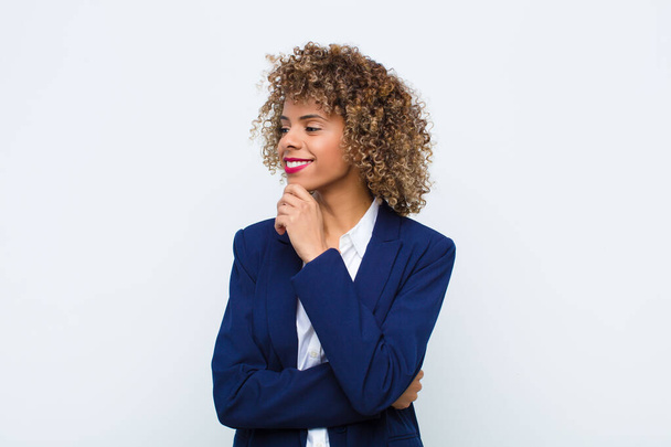 young woman african american smiling with a happy, confident expression with hand on chin, wondering and looking to the side against flat wall - Foto, Imagem