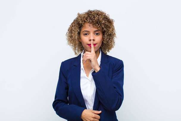 joven mujer afroamericana mirada seria y cruz con el dedo presionado a los labios exigiendo silencio o silencio, manteniendo un secreto contra la pared plana
 - Foto, imagen