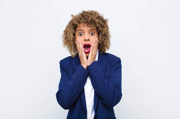 young woman african american feeling shocked and scared, looking terrified with open mouth and hands on cheeks against flat wall - Foto, Bild