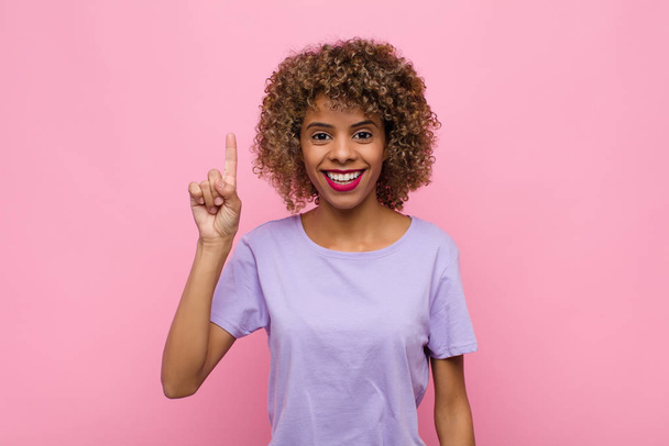 young african american woman smiling cheerfully and happily, pointing upwards with one hand to copy space against pink wall - 写真・画像