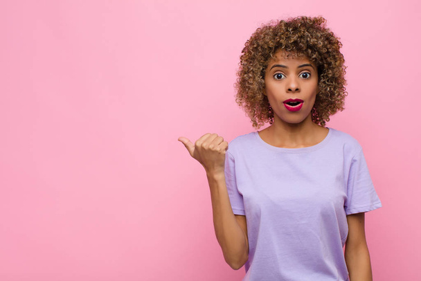 young african american woman looking astonished in disbelief, pointing at object on the side and saying wow, unbelievable against pink wall - Fotoğraf, Görsel