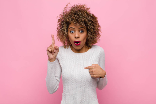mladá afroamerická žena se cítí hrdá a překvapená, ukazuje na sebe sebevědomě, cítí se jako úspěšná jednička na růžové stěně - Fotografie, Obrázek