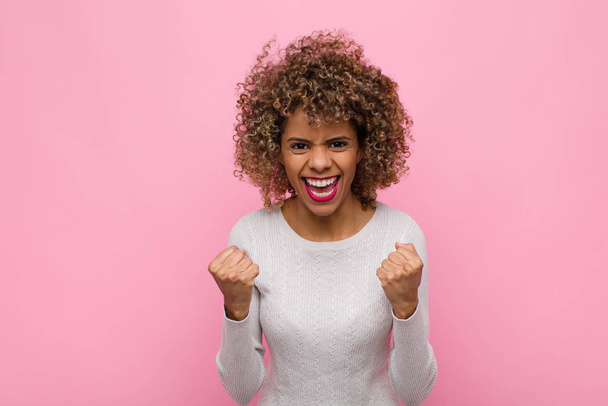 joven mujer afroamericana gritando triunfalmente, riendo y sintiéndose feliz y emocionada mientras celebra el éxito contra la pared rosa
 - Foto, imagen