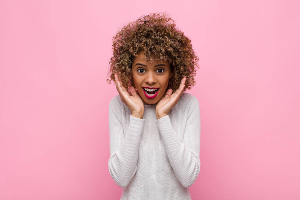 young african american woman feeling shocked and excited, laughing, amazed and happy because of an unexpected surprise against pink wall - Foto, afbeelding