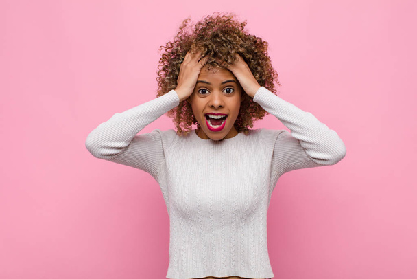 young african american woman raising hands to head, open-mouthed, feeling extremely lucky, surprised, excited and happy against pink wall - Foto, imagen