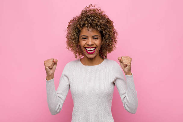 young african american woman feeling happy, positive and successful, celebrating victory, achievements or good luck against pink wall - Foto, afbeelding
