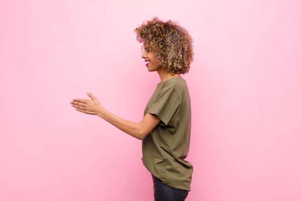 young african american woman smiling, greeting you and offering a hand shake to close a successful deal, cooperation concept against pink wall - Foto, Bild