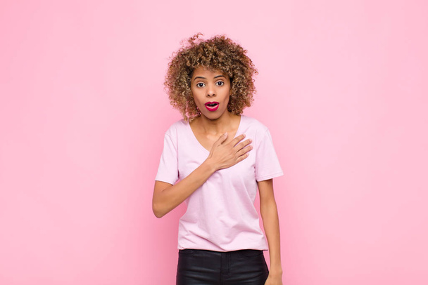 young african american woman feeling shocked and surprised, smiling, taking hand to heart, happy to be the one or showing gratitude against pink wall - 写真・画像