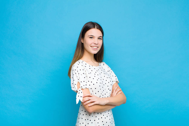 young pretty woman smiling to camera with crossed arms and a happy, confident, satisfied expression, lateral view against blue wall - Foto, Imagem
