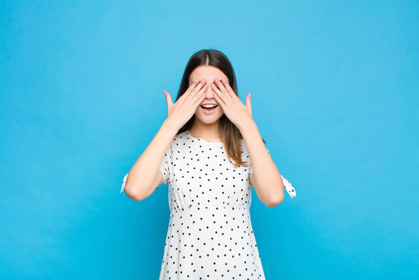 young pretty woman smiling and feeling happy, covering eyes with both hands and waiting for unbelievable surprise against blue wall - Fotoğraf, Görsel